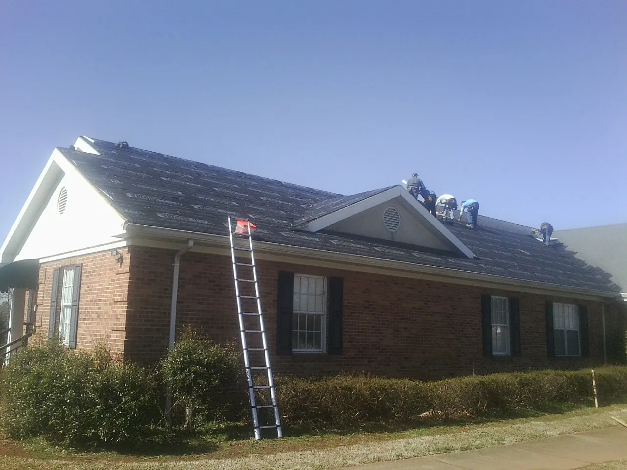 Crew tearing shingles off a brick ranch roof with a ladder set — Emergency Roof Repair work by our crew in Romney