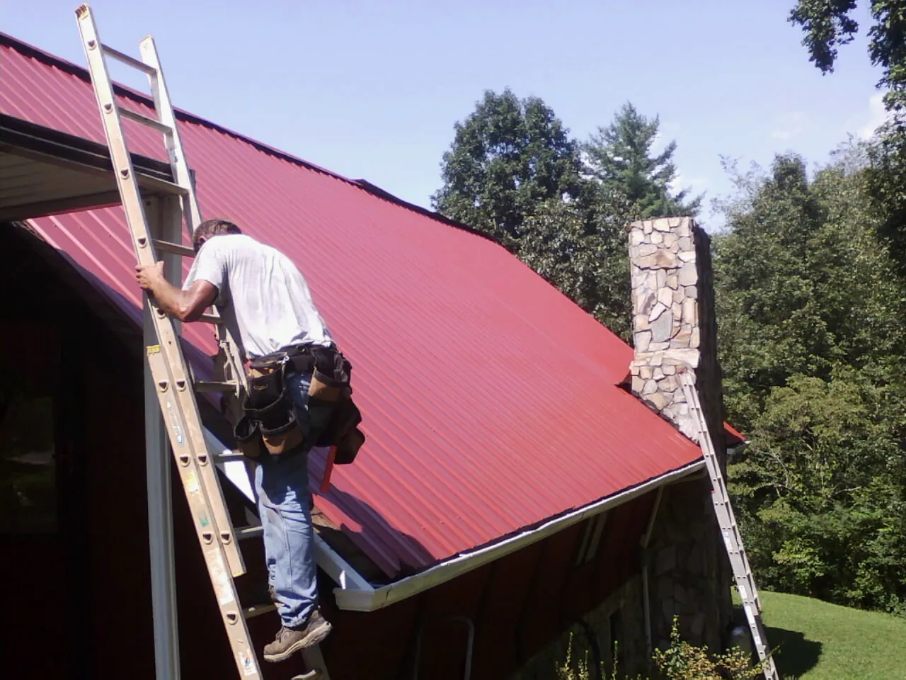 Roofer on a ladder beside a red metal roof and stone chimney — Soffit & Fascia Repair by Roofing Brothers in Romney